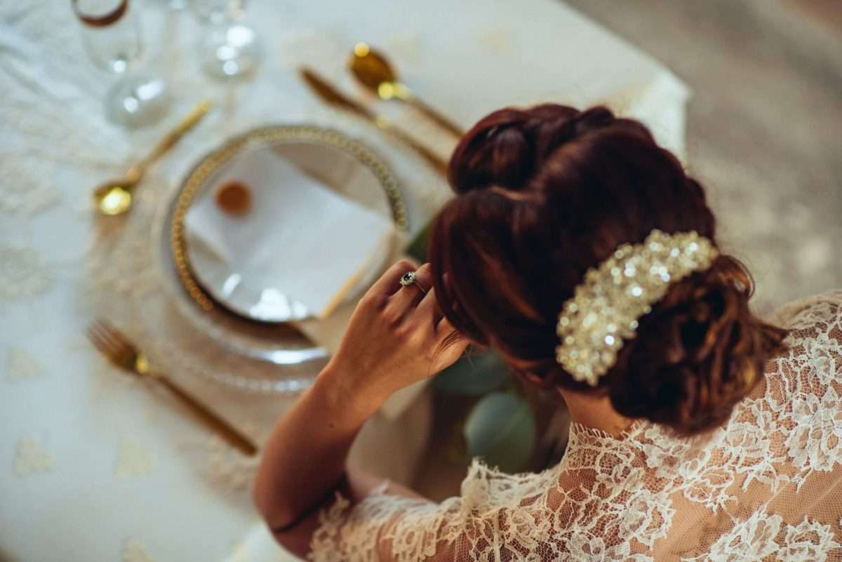 Foto artistica di sposa seduta a tavola imbandita per il matrimonio. Abito da sposa in pizzo bianco e ferma capelli in diamanti