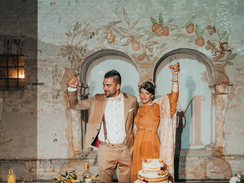 Foto degli sposi al momento del taglio torta, nei loro vestiti sartoriali, con una proiezione nello sfondo del loro monogramma che si fonde perfettamente con i due archi presenti nel muro.